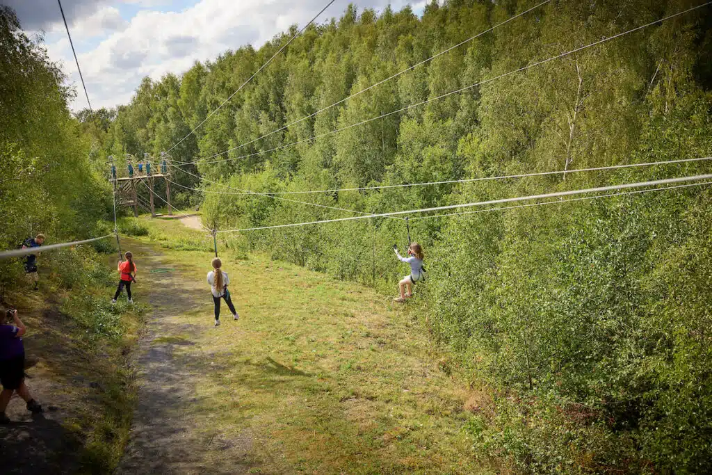 Several people ride zip lines over a grassy area surrounded by trees, with others watching from the ground and a platform in the background.