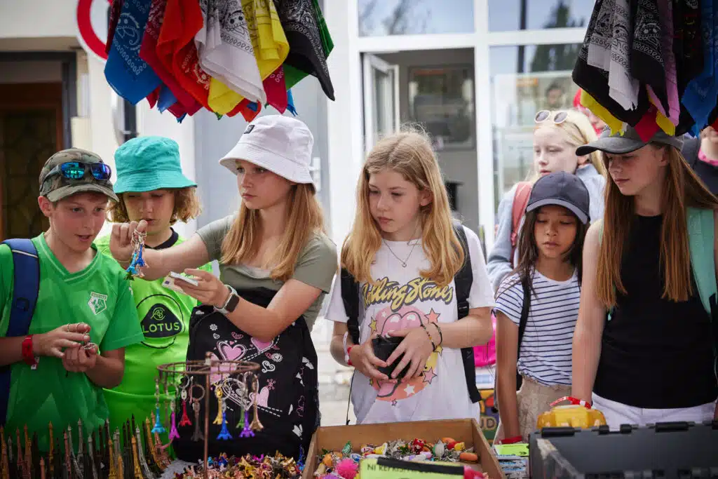 A group of children stands at a multi-activity outdoor market stall, examining small items and souvenirs displayed on tables under hanging colorful cloths.