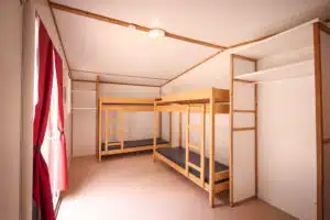 A clean, sparsely furnished room with two wooden bunk beds, bare mattresses, an open wardrobe, and red curtains on a window.