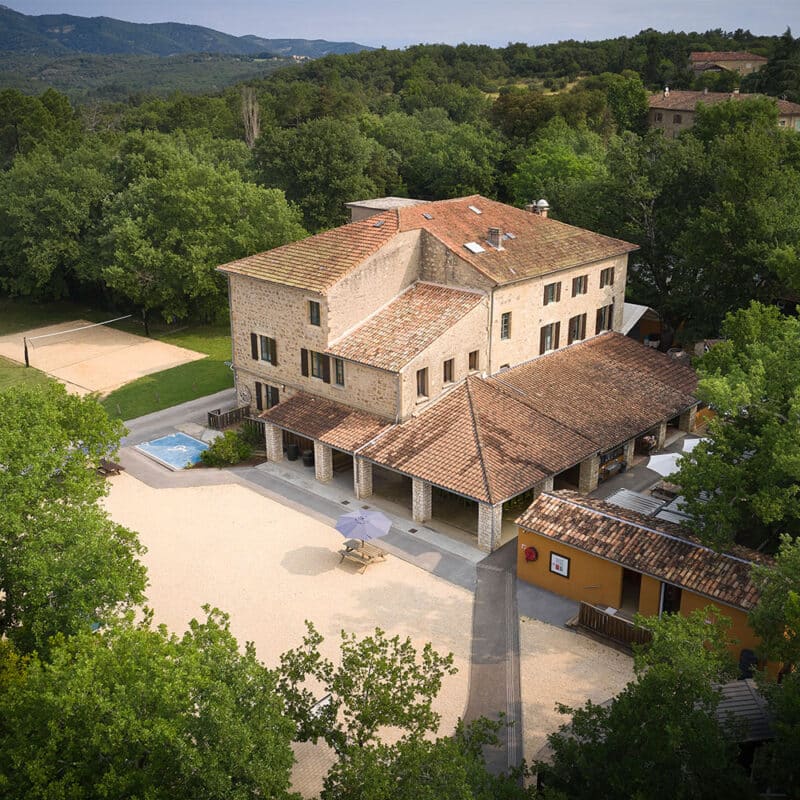 A large stone building with terracotta roofs is surrounded by trees, with a volleyball court, small pool, and outdoor seating area visible in the courtyard.