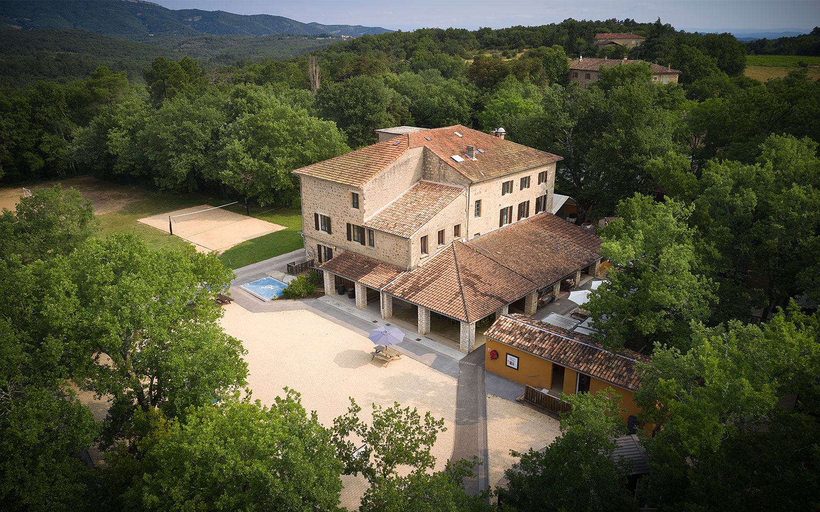 A large stone building with terracotta roofs is surrounded by trees, with a volleyball court, small pool, and outdoor seating area visible in the courtyard.