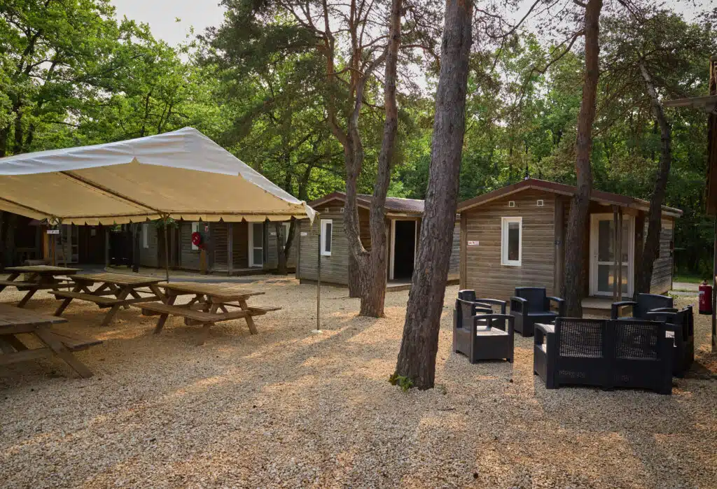 Outdoor campsite area with wooden cabins, plastic chairs around a small table, picnic tables under a canopy, and trees providing shade.