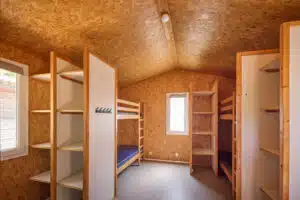 Room with wood-paneled walls and ceiling, two sets of bunk beds, open shelving units, and windows letting in natural light.