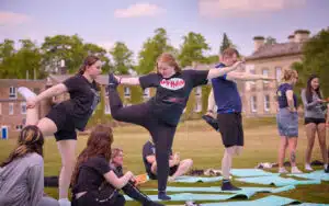 A group of people practice stretching and balancing exercises on a grassy field, using yoga mats, with buildings in the background—a perfect spot for a multi-activity outdoor session.