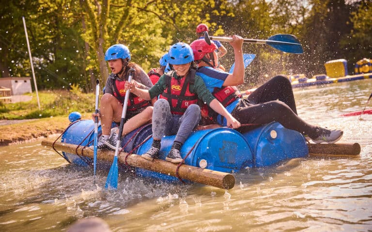 Four people wearing helmets and life jackets paddle a makeshift raft of barrels and logs on a small body of water, splashing as they move forward during this fun multi-activity adventure.
