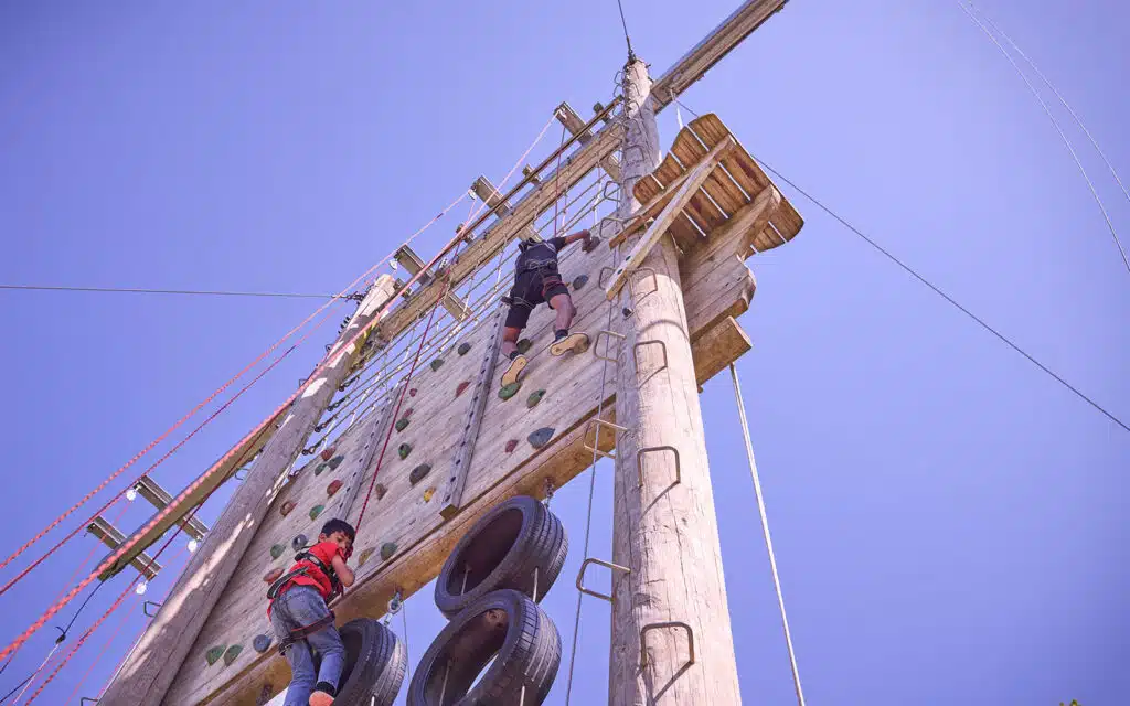 Two people wearing harnesses climb a tall outdoor multi-activity wooden wall, featuring ropes and tires, under a clear blue sky.