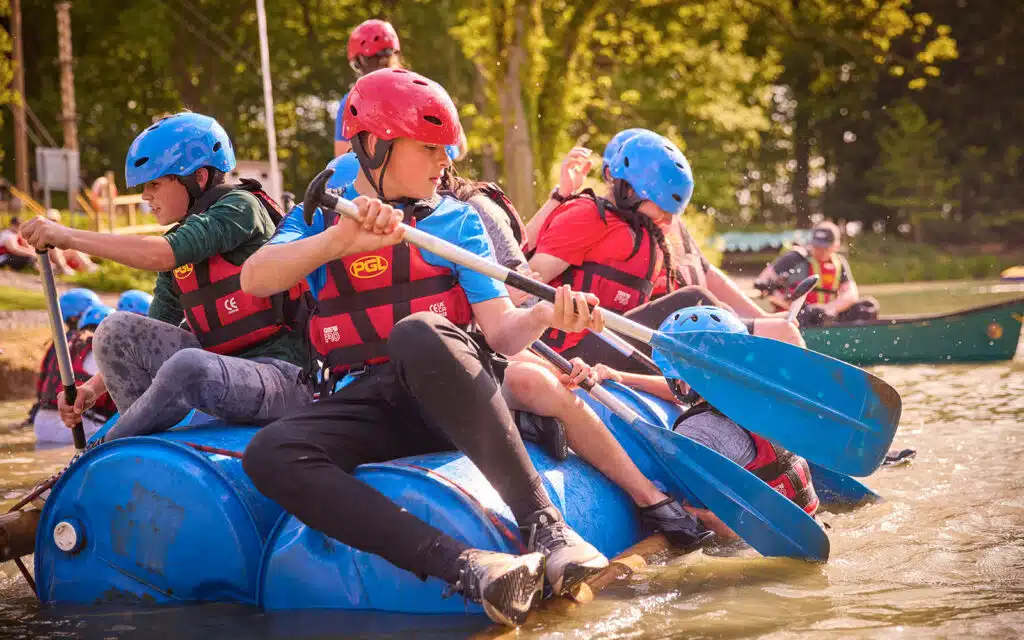 A group of young people wearing helmets and life jackets paddle a makeshift raft made of blue barrels on a lake during a multi-activity adventure.