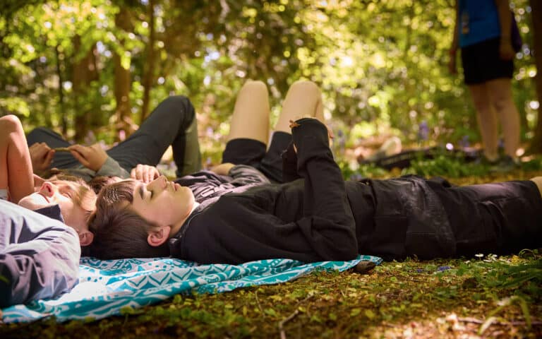 Several people lie on a blanket in a sunlit forest clearing, enjoying a multi-activity day as they relax and rest, surrounded by trees and greenery.