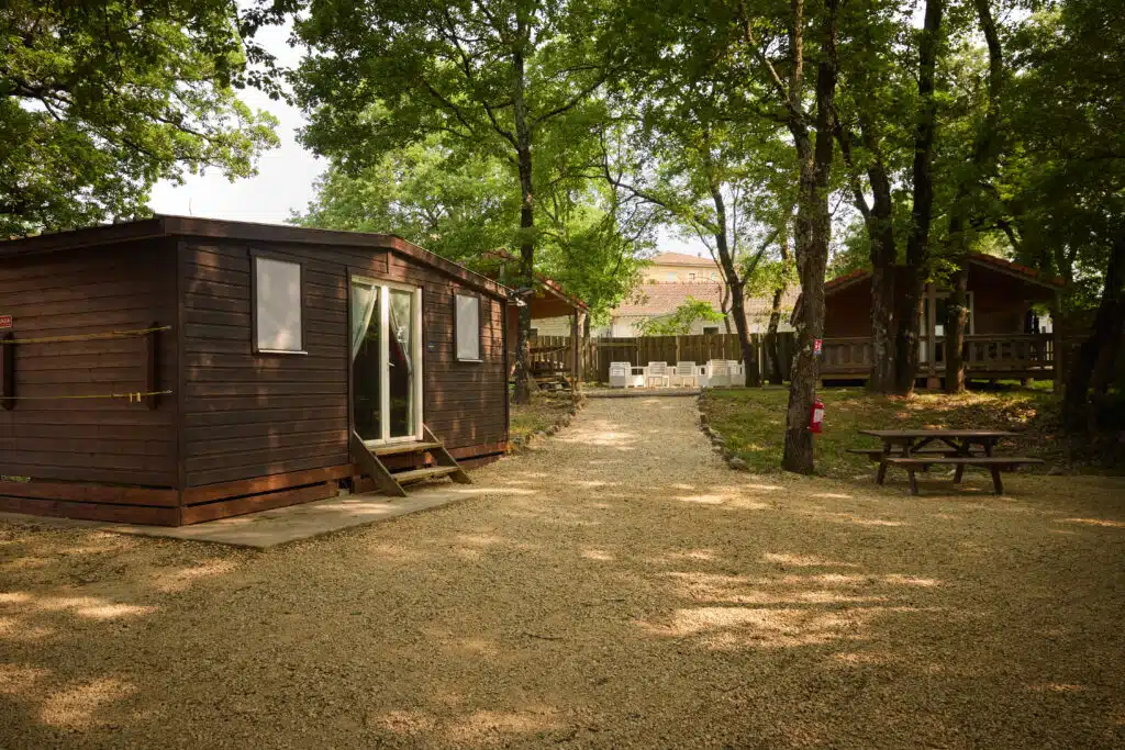 Small wooden cabin and picnic table on a gravel area surrounded by trees, with more buildings and outdoor seating visible in the background.