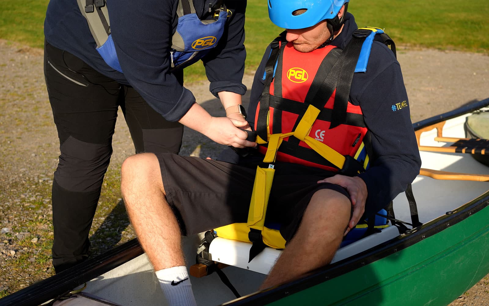 A person sitting in a canoe has their life jacket straps checked and secured by another individual. Both are wearing safety gear, including helmets and life vests.