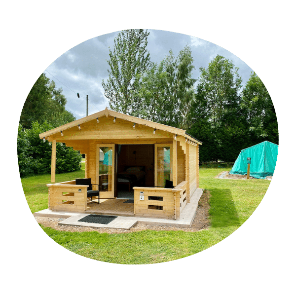 Small wooden cabin with a covered porch sits on a grassy area, surrounded by trees, with a covered structure visible nearby under a cloudy sky.