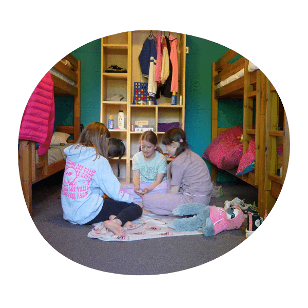 Four girls sit on a blanket on the floor of a cabin dorm room with bunk beds, talking and playing together. Shelves with clothes and personal items are visible in the background.