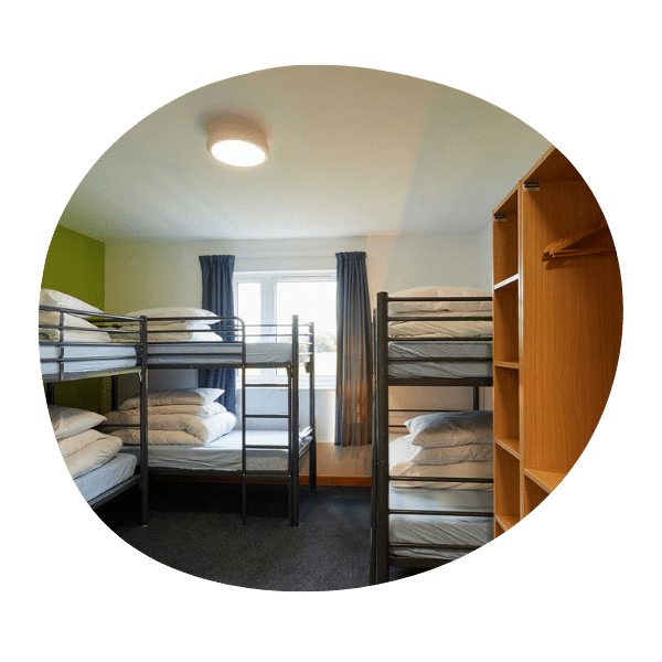 A room with two metal bunk beds, each with white bedding, a window with blue curtains, and an open wooden shelving unit on the right side.