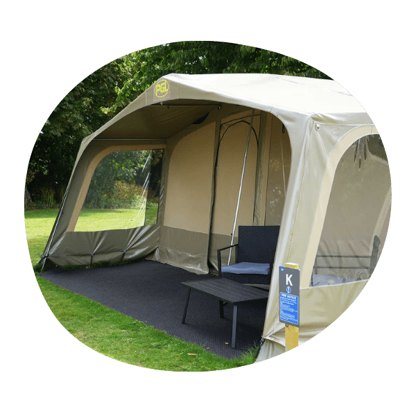 A beige outdoor tent with clear side panels, containing a black bench and table, set on a grassy area with trees in the background.