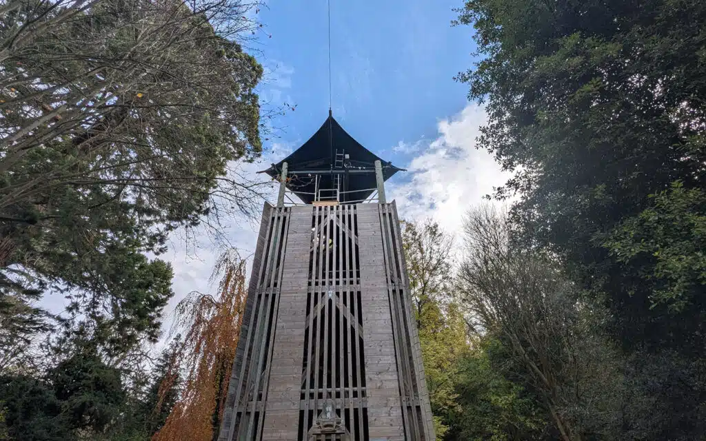 A tall wooden observation tower with a black roof stands among trees under a partly cloudy sky, viewed from below.