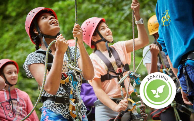 A group of children wearing helmets and harnesses participate in an outdoor climbing activity; a badge labeled "Adapting" is visible in the foreground.