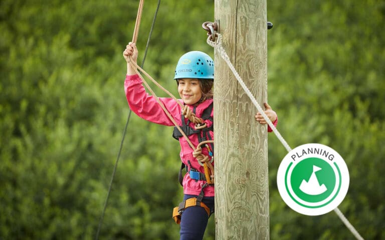 A child wearing a helmet and harness holds onto a wooden pole and rope outdoors, with a "Planning" badge icon in the corner.