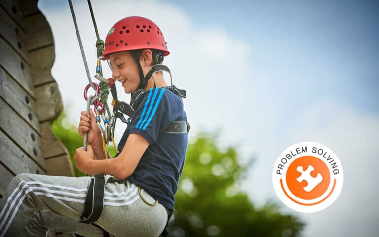 A child wearing a helmet and harness climbs a ropes course outdoors, smiling. A "problem solving" badge with a puzzle piece icon is shown in the corner.