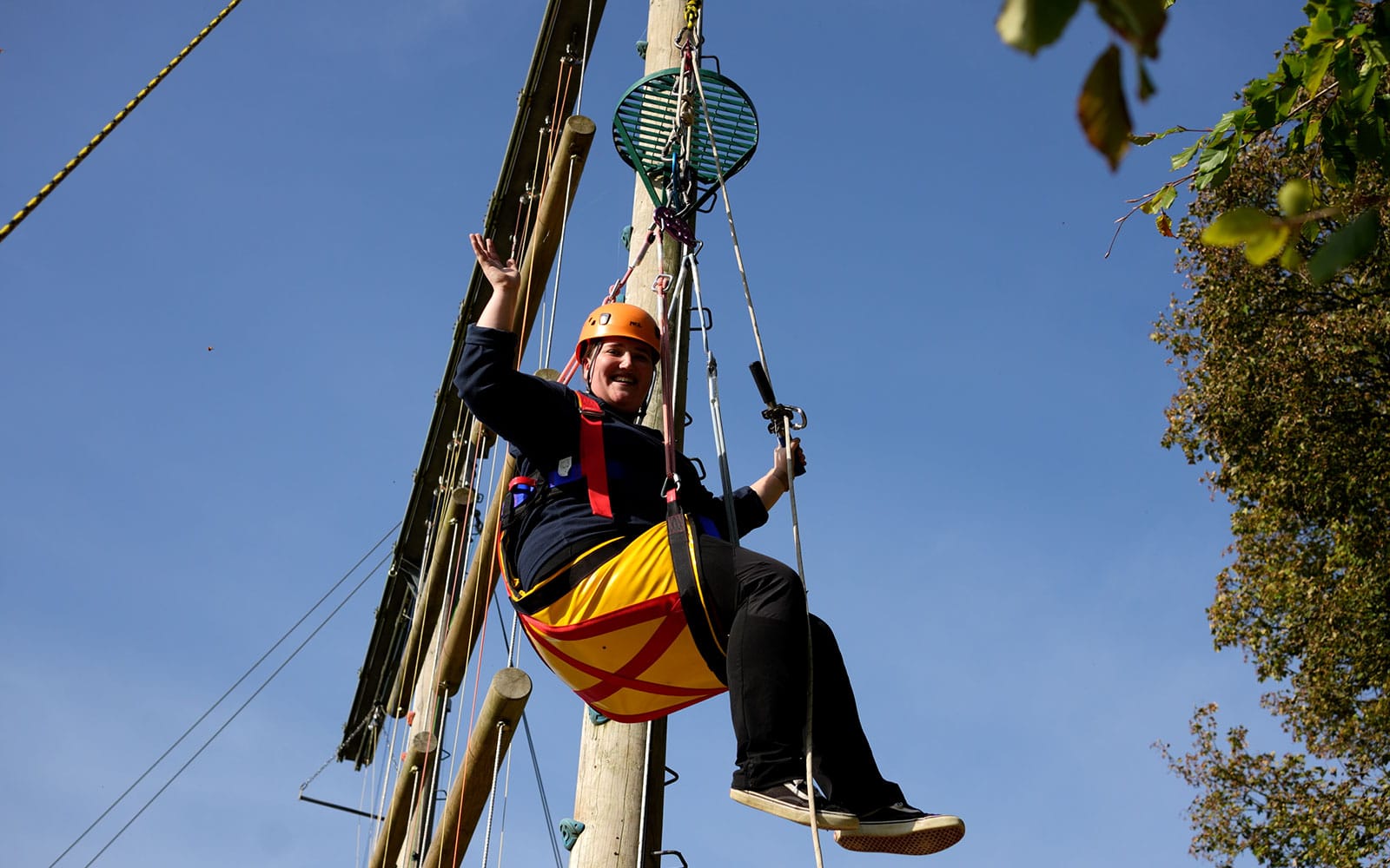 A person wearing a helmet and harness sits in a suspended swing, waving with one hand, against a clear blue sky and climbing structure.
