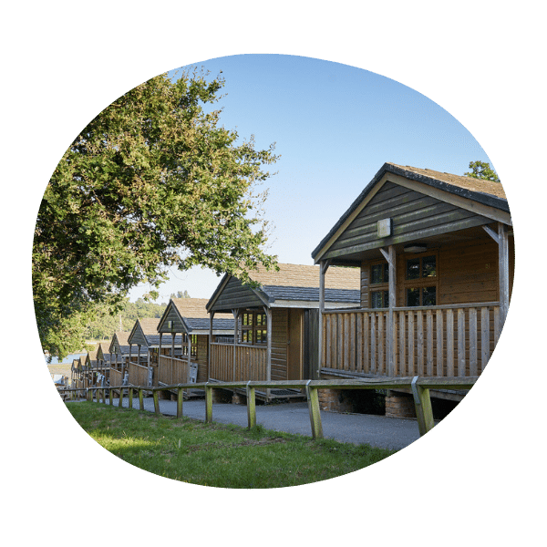 Row of wooden cabins with front porches, set along a pathway and shaded by trees on a sunny day.