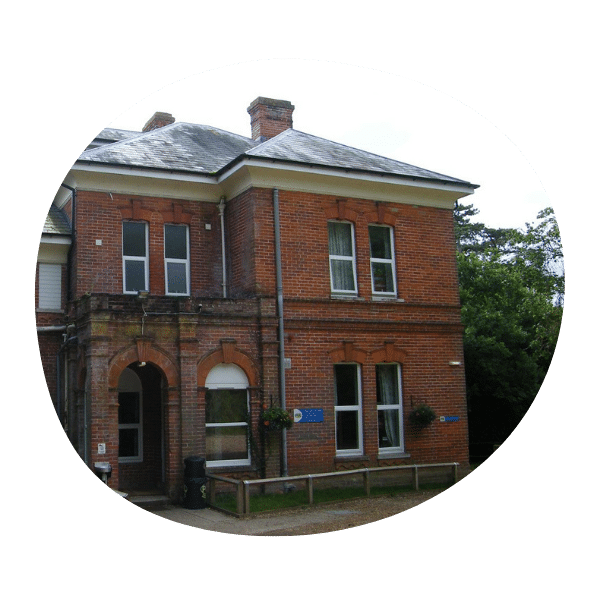 A two-story red brick building with white trim, arched entrance, and rectangular windows, surrounded by greenery.