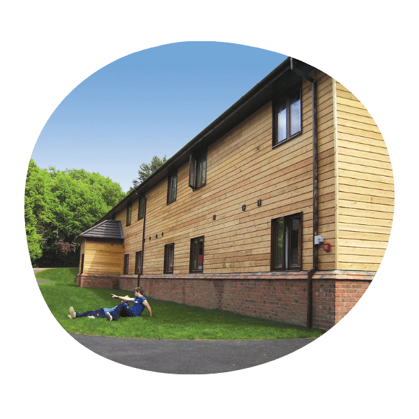 A person wearing a blue shirt and jeans lies on the grass outside a two-story wooden building under a clear blue sky.