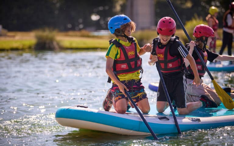Three children wearing helmets and life jackets smile and paddle while kneeling on a paddleboard on a sunny lake.