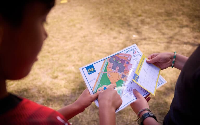 Two people hold and examine a detailed map and a small card outdoors, possibly preparing for orienteering or a similar activity.