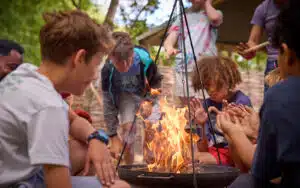 A group of children sit and stand around an outdoor fire pit, warming their hands and watching the flames.