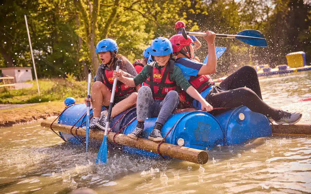 Four people wearing helmets and life vests paddle a homemade raft made of blue barrels, wooden poles, and rope on a calm outdoor body of water, splashing as they move.