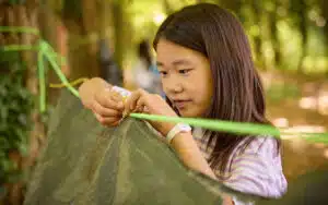 A girl outdoors is tying a green rope to secure a tarp between trees, focusing intently on her task.