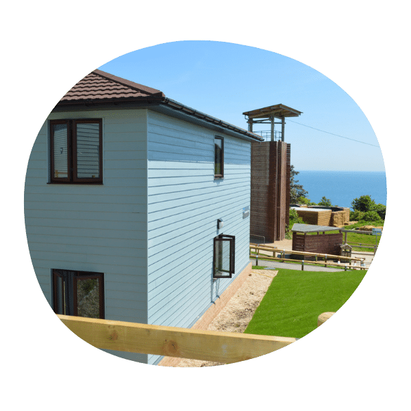 A modern light blue two-story house with a brown roof overlooks a green lawn and the sea in the background on a clear day.