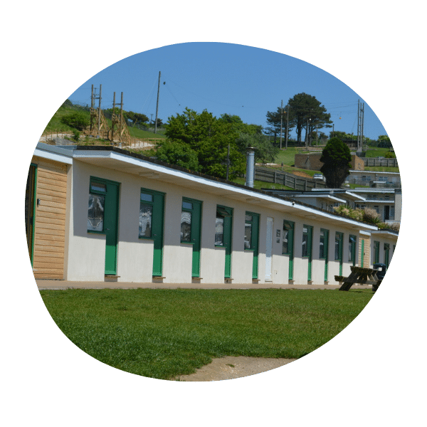 A single-story building with green doors and windows sits on a grassy area, with trees and construction visible in the background under a clear blue sky.
