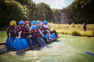 A group of children wearing helmets and life vests sit on a makeshift raft made of barrels and logs, floating on a lake near a grassy shore and wooden tower.