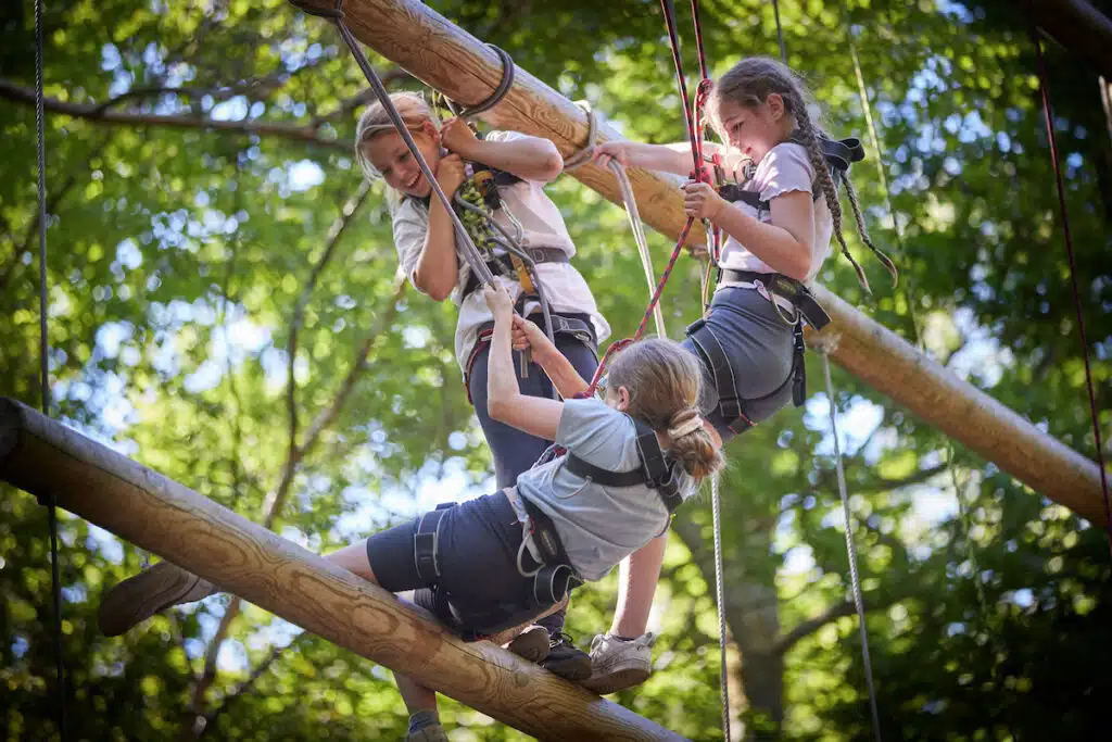 Three children in harnesses climb on wooden beams suspended outdoors, surrounded by green foliage, during a ropes course activity.