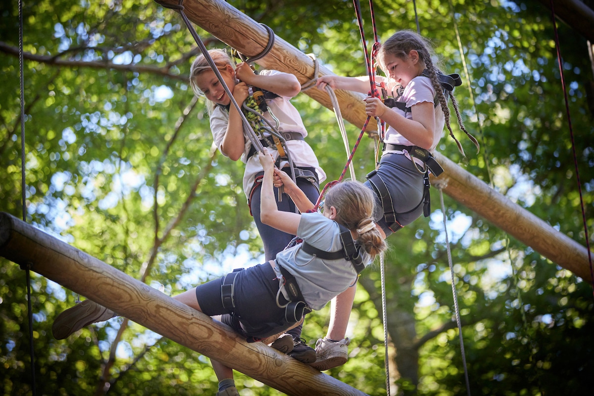 Three children in harnesses climb on wooden beams suspended outdoors, surrounded by green foliage, during a ropes course activity.