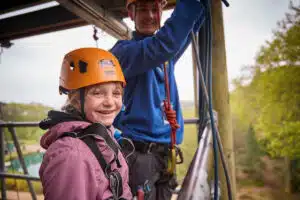 A young girl wearing a helmet and harness smiles at the camera while standing next to an instructor on a zip line platform outdoors.