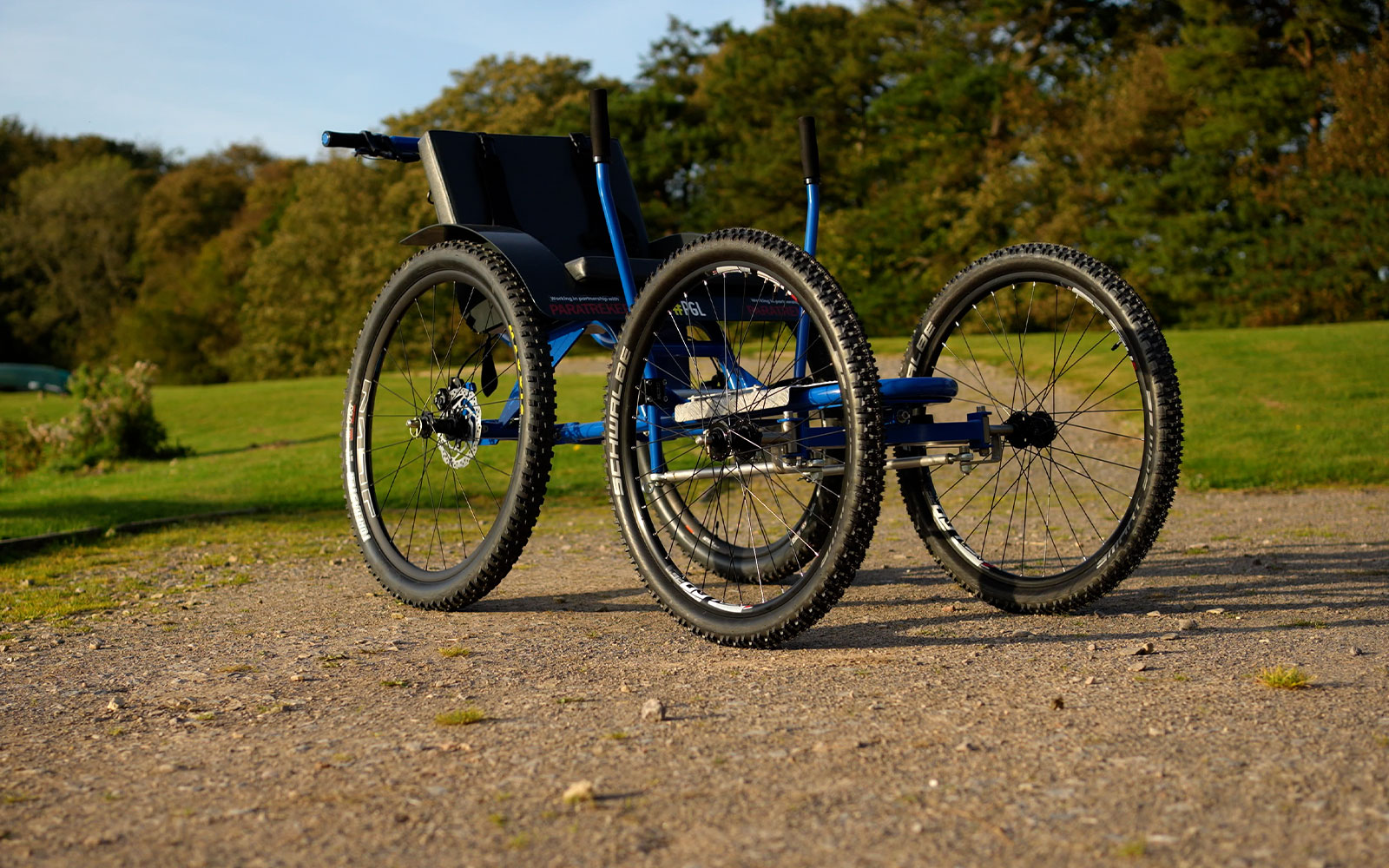 A blue off-road wheelchair with large, rugged tires is positioned on a gravel path with grass and trees in the background.
