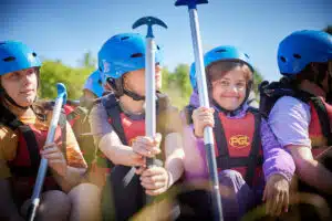 A group of children in blue helmets and red life jackets hold paddles while sitting outdoors, preparing for a water activity.