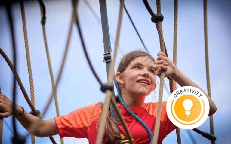 A girl in an orange shirt climbs a multi-activity rope structure outdoors, looking upward. A “Creativity” badge with a lightbulb icon appears in the lower right.
