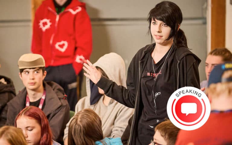 A young person stands and speaks to a seated group of peers in a casual indoor setting; a "speaking" icon appears in the foreground, highlighting this multi-activity space designed for interactive discussions.