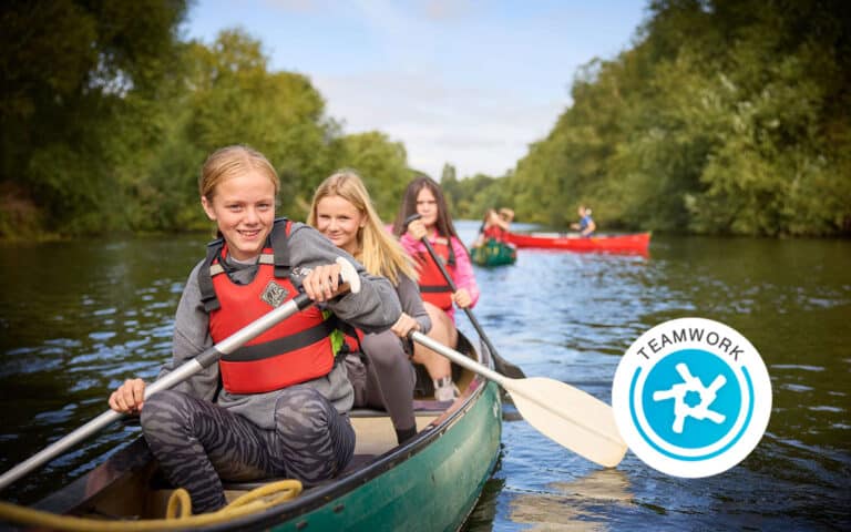 Four girls wearing life jackets paddle canoes on a river surrounded by trees, showcasing the spirit of teamwork during this multi-activity adventure. A "Teamwork" badge graphic appears in the bottom right corner.