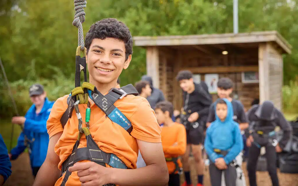 A boy wearing a harness and orange shirt smiles at the camera during a multi-activity day, with other people and a wooden shelter blurred in the background.