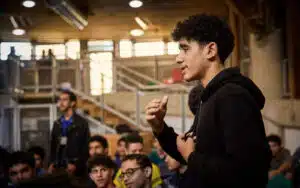 A young man in a black hoodie stands and speaks in front of a seated group of people in an indoor, multi-activity space with exposed beams and metal railings.