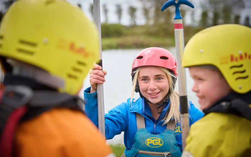 A woman wearing a pink helmet and blue jacket talks to two children in yellow helmets and life jackets near a body of water, all holding paddles.