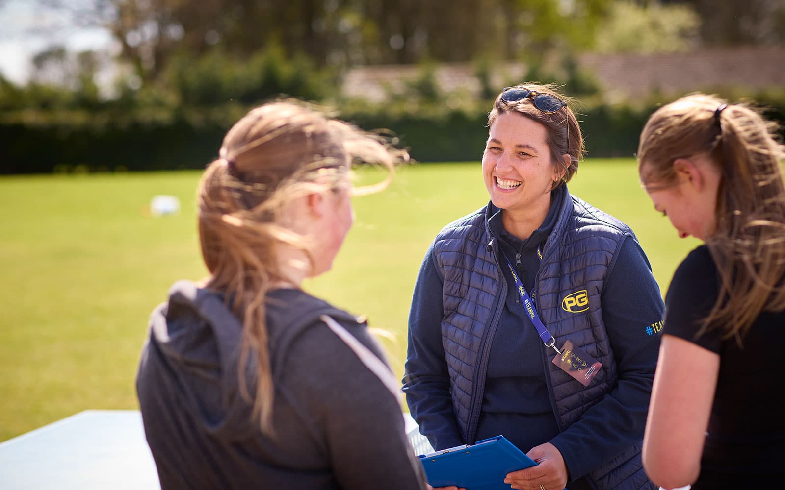 A woman wearing a blue vest and sunglasses talks with two girls outdoors on a sunny day, holding a clipboard.