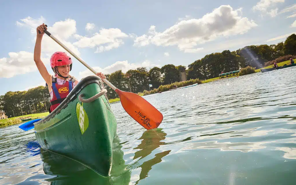 A person wearing a helmet and life vest paddles a green canoe on a calm lake under a partly cloudy sky.