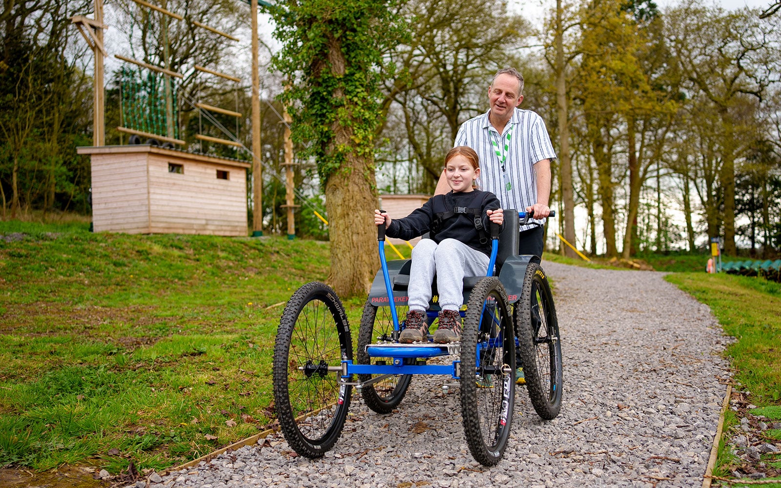 A man pushes a girl in an all-terrain wheelchair along a gravel path in a park, with trees and a wooden building in the background.