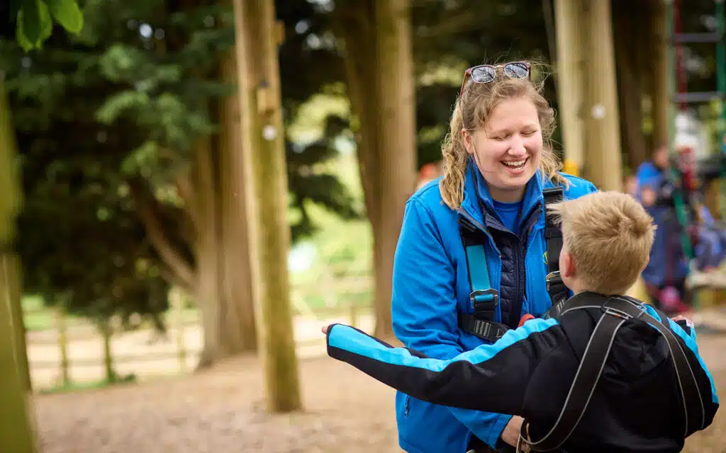 An adult and a child wearing blue jackets and harnesses smile and interact outdoors near wooden poles and trees, likely at an adventure park or ropes course.