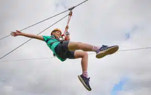 A person wearing a helmet and harness rides a zipline against a cloudy sky, with arms and legs outstretched.